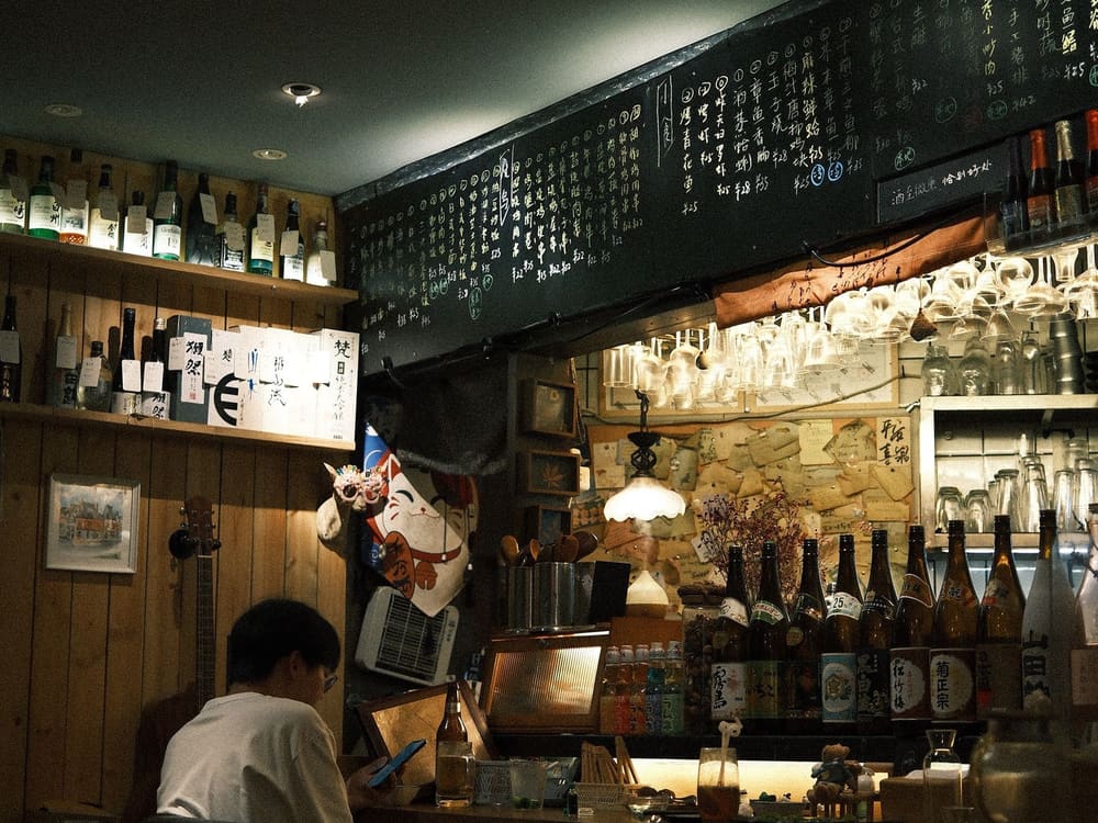 酒瓶が並ぶ棚と、カウンターの中に立つ人。暖色の灯りの夜の店内